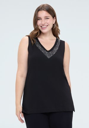 Glunderende vrouw met golvend bruin haar draagt een mouwloze zwarte top met steendetail rond de V-hals tegen een lichtgrijze achtergrond.