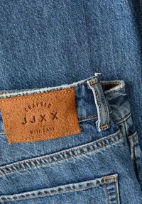 Plave traperice s smešnim kožnim aplikacijom označenim "CRAFTED JJXX WITH CARE", vidljivim šavovima i blago istrošenom teksturom.