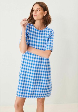 Kvinde med brunt hår iført blå og hvid ternet kjole med korte ærmer, står med armene krydset og ser til siden.