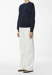 Mörkblå sweatshirt med rundad halsringning och ribbade mudd, tillsammans med vida vita byxor och svarta sandaler, vilket skapar en avslappnad look.