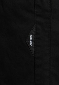 Tela negra con una textura suave. Presenta una etiqueta en forma de diamante con "CHASIN'" en costura blanca. Diseño simple, sin patrones adicionales.