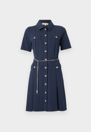 Robe chemise bleu marine à manches courtes avec boutons argentés, quatre poches à rabat, et une ceinture-chaîne argentée suspendue lâchement à la taille.