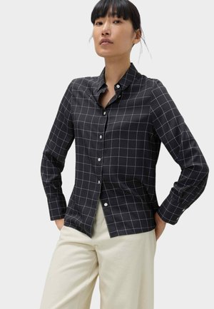 Kvinde iført sort langærmet skjorte med knapper og hvidternet mønster samt cremefarvede bukser med vide ben, hænderne i lommerne.