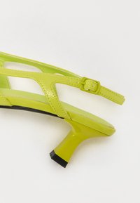Sandale à talons en cuir verni vert avec des brides entrecroisées et une boucle réglable, présentant un talon évasé élégant et une surface texturée.