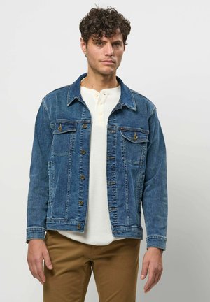 Mand med krøllet hår iført en knappet blå denimjakke over en hvid henley-trøje og brune bukser mod en ensfarvet baggrund.