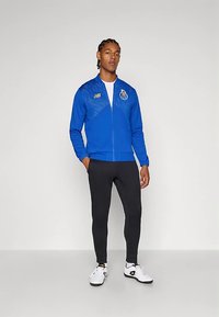 Blauer Reißverschluss-Pullover mit strukturiertem Design und Team-Logo, kombiniert mit schwarzen, eng anliegenden Hosen und weißen Sportschuhen.