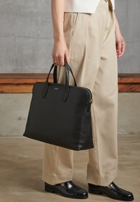 Borsa a mano in pelle nera con doppio manico, texture liscia, forma rettangolare e delicato dettaglio del logo sul davanti. Indossata con pantaloni beige.
