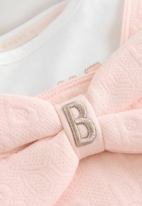 Vaaleanpunainen kangastoppi, jossa on suuri muotoinen solmu ja hopeinen brodeerattu "B"-kirjain. Valkoinen kaulus alapuolella, jossa on hienovarainen brändäys.