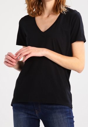 Woman wearing a plain black V-neck short-sleeve shirt and blue jeans, with hands gently clasped in front.