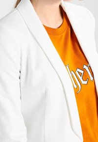 Witte blazer met een gelapelde revers, gedragen over een oranje T-shirt. De blazer heeft een gladde textuur en een aansluitend ontwerp.