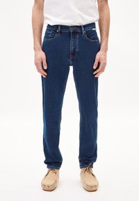 Vaqueros de mezclilla oscuros con un corte Slim, diseño de cinco bolsillos, detalles de costura visibles y una sección sutilmente descolorida en la parte delantera.