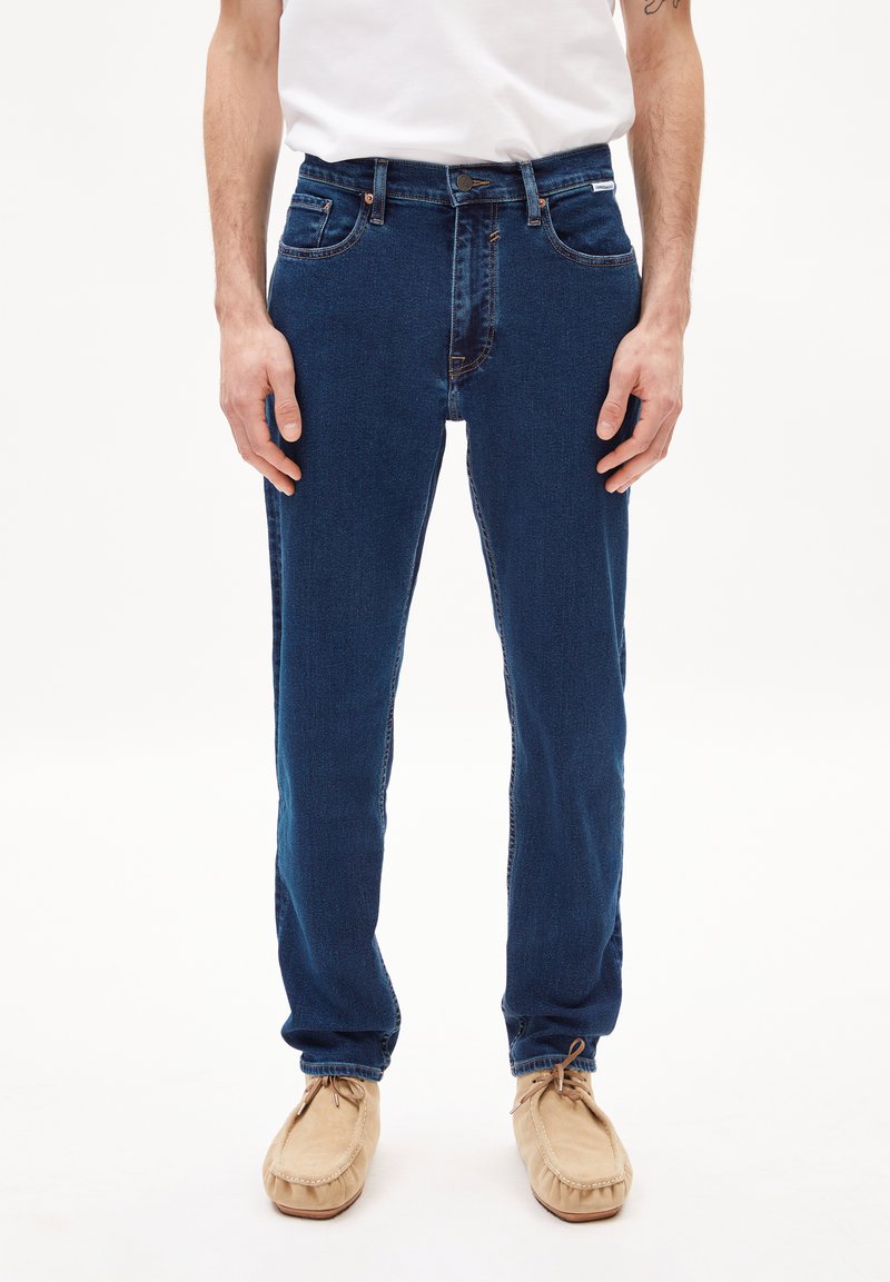 Vaqueros de mezclilla oscuros con un corte Slim, diseño de cinco bolsillos, detalles de costura visibles y una sección sutilmente descolorida en la parte delantera.