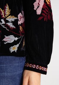 Haut en velours noir avec broderie florale en rose, blanc et jaune. Présente un poignet de manche froncé avec un bord à motifs.