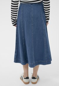 Persoon in een blauwe denim midi rok, een zwart-wit gestreepte trui en witte sneakers met zwarte accenten, staande tegen een witte achtergrond.
