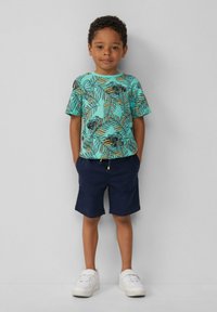 T-shirt corta turchese con motivo di foglie di palma nere e arancioni, abbinata a pantaloni corti blu navy. Sneakers bianche completano l'outfit.