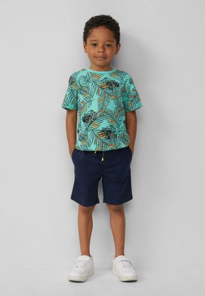 Turquoise T-shirt met korte mouwen, bedrukt met een zwart en oranje palmenbladpatroon, gecombineerd met marineblauwe shorts. Witte sneakers maken de outfit af.