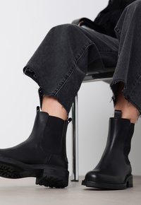 Botas negras de cuero hasta el tobillo con paneles laterales elásticos, suelas de goma texturizadas y tiradores; combinadas con unos vaqueros de mezclilla negros deshilachados.