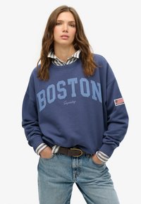 Sudadera azul con "BOSTON" en un texto más grande, puños acanalados y un pequeño parche de la bandera americana en la manga derecha sobre una camisa a rayas.
