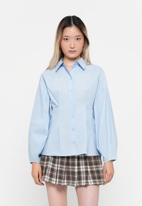 Camicia azzurra chiara con colletto, maniche lunghe a palloncino e vita aderente. Abbinata a una gonna a pieghe in tartan.