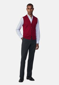 Gilet rosso lavorato a maglia con scollo a V e bottoni indossato sopra una camicia colletto blu chiaro, abbinato a pantaloni grigio scuro e scarpe nere.