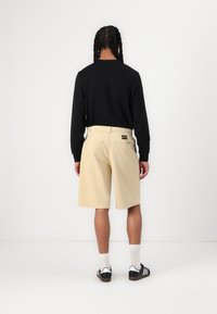 Beige bomuldsshorts med en afslappet pasform, der har en baglomme og et sort logo patch. Sammenstylet med en sort langærmet skjorte og sneakers.