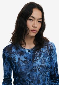 Gilet bleu à motifs avec un col rond, fermeture à boutons et manches longues. Présente un motif floral et un tissu texturé.