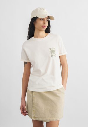 Femeie purtând o șapcă bej, tricou alb cu imprimeu "Roxy 1990" și fustă bej, stând cu o mână în buzunar.