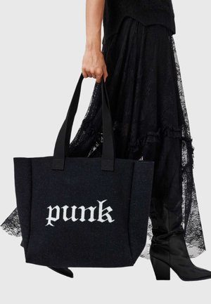 Μαύρη τσάντα με στυλ tote με λευκό κείμενο "punk", κατασκευασμένη από υλικό με υφή. Διαθέτει δύο ανθεκτικές λαβές και δομημένο σχήμα, σε συνδυασμό με μια φούστα δαντέλας.