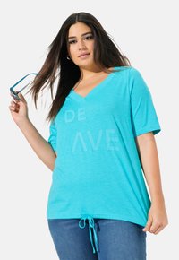 Una camiseta de cuello en V de manga corta en color turquesa, con un dobladillo ajustable y un diseño de texto impreso. La tela parece ligera y texturizada.