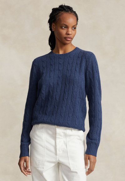 Naine, kes kannab sinist palmikutrikootud kampsunit ja valgeid pükse, seisab beeži ühevärvilise tausta ees ning vaatab vasakule.