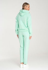 Miętowy bluza z kapturem i dopasowane spodnie dresowe z miękkiego materiału. Bluza ma dużą tylną kieszeń i sznurki, a spodnie mają dopasowane mankiety. Białe buty.