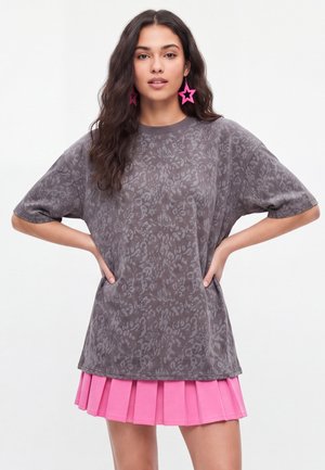 Kvinde med langt mørkt hår bærer oversized grå mønstret T-shirt, rosa plisseret nederdel og store lyserøde stjerneformede øreringe med hænderne på hofterne.