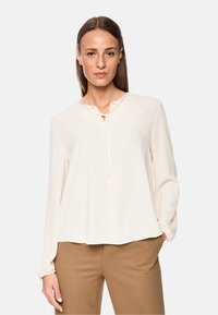 Helle beige Bluse mit lockerer Passform, V-Ausschnitt und Kordelzugverschluss. Lange Ärmel mit gerafften Bündchen. Glatte, weiche Stofftextur.
