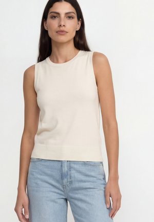 Femme aux longs cheveux foncés portant un haut crème sans manches et un jean bleu clair, debout devant un fond blanc uni.