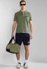 Polo shirt verde oliva, pantaloni corti blu navy e sneakers bianchi; mentre porta una borsa a tracolla verde e nera, posato su uno sfondo neutro.