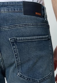 Gros plan sur la poche arrière d'un jean en denim bleu avec une étiquette noire affichant le logo orange "BOSS" sur la ceinture.