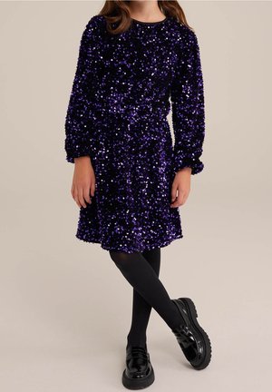 Enfant portant une robe à sequins violette à manches longues, des collants noirs et des mocassins noirs brillants, debout jambes croisées.
