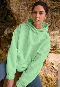 Fuzzy green oversized hoodie with a hood, ribbed cuffs, and a soft texture, worn with blue leggings in a natural outdoor setting.