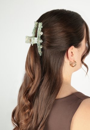 Cheveux bruns ondulés mi-attachés avec une pince à cheveux florale verte translucide, porte une boucle d'oreille créole dorée et un haut marron sans manches sur fond blanc.
