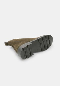 Botină din suede maro, cu o talpă din cauciuc texturat negru, având vârful rotunjit și o buclă pe spate pentru o purtare ușoară.