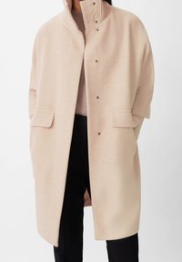 Beige villasekoite takki, jossa korkea kaulus, nepparikiinnitys ja sivutaskut; siinä on väljä istuvuus ja pehmeä tekstuuri. Sopii kerrospukeutumiseen.