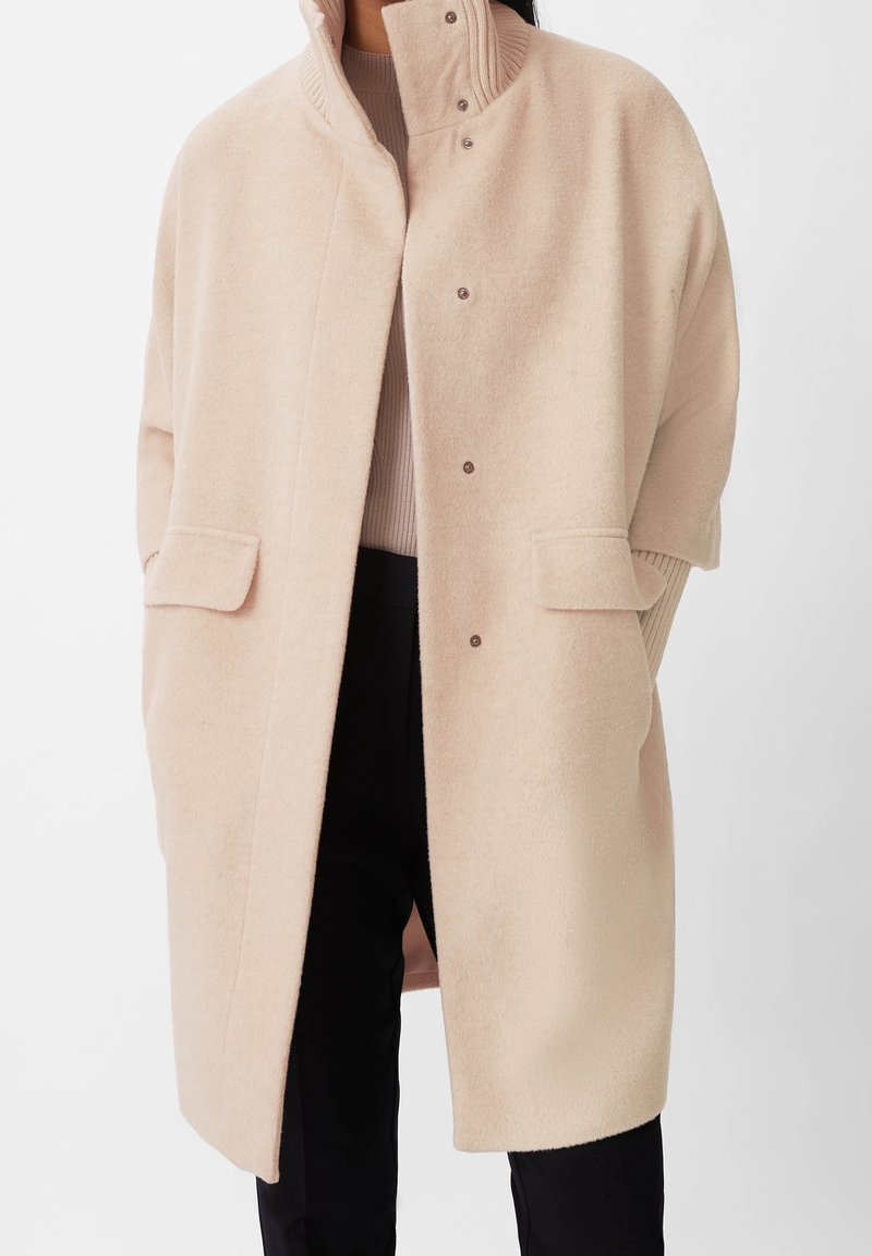 Beige villasekoite takki, jossa korkea kaulus, nepparikiinnitys ja sivutaskut; siinä on väljä istuvuus ja pehmeä tekstuuri. Sopii kerrospukeutumiseen.