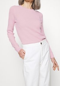 Kvinna som bär en ljusrosa texturerad långärmad topp nedstoppa i högmidjade vita byxor med en hand i fickan.