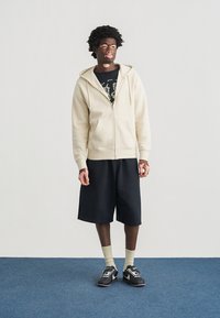 Μπεζ φούτερ με φερμουάρ, μαύρο μπλουζάκι με στάμπα, μαύρο σορτς, ανοιχτόχρωμες κάλτσες και μαύρα αθλητικά παπούτσια. Το μοντέλο στέκεται πάνω σε μπλε χαλί μπροστά σε απλό φόντο.