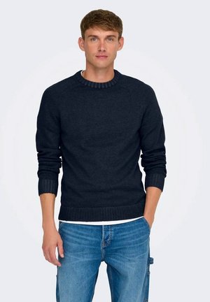 Pullover in maglia blu navy scuro con collo a giro, polsini e orlo a costine, indossato sopra una camicia bianca, abbinato a jeans in denim blu.