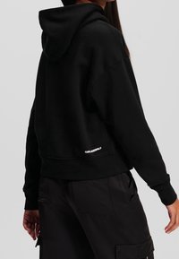 Czarna krótka bluza z kapturem ze ściągaczem, luźnym krojem, prążkowanymi mankietami i dołem. Na lewej dolnej stronie znajduje się biały logotyp.