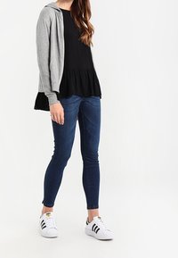 Felpa grigia, top peplum nero, jeans aderenti blu e sneakers bianche con strisce nere. Tessuto testurizzato e stile casual.