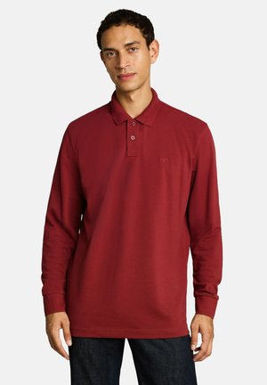Man wearing a long-sleeve maroon polo shirt with buttoned collar and dark pants, standing against a plain light background.