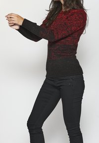 Gestrickter Pullover in Rot und Schwarz mit strukturiertem Muster, ausgestattet mit rundem Ausschnitt und gerippten Bündchen. Kombiniert mit eng anliegenden schwarzen Jeans.