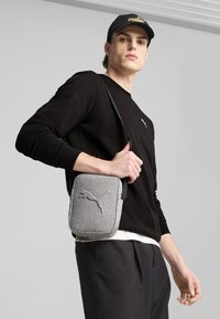 Borsa a tracolla grigia in tessuto con logo Puma, forma rettangolare, tracolla regolabile e superficie testurizzata, indossata da un modello in abbigliamento casual.